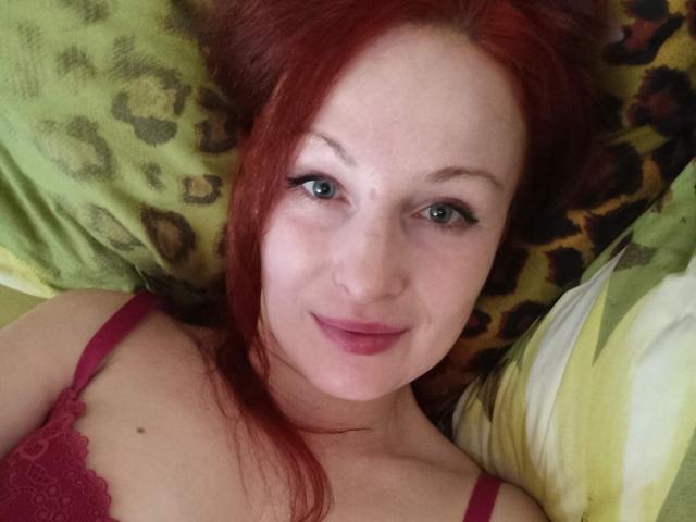 MarleneDavies with red hair, green eyes, and pink lipstick, wearing a red lace bra, lying on a leopard print pillow, smiling at the camera.