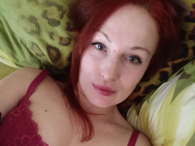 Marlene Davies, com cabelo ruivo, olhos verdes e batom rosa, deitada em um travesseiro com estampa de leopardo, vestindo um sutiã de renda vermelho, sorrindo suavemente. Seus seios estão ligeiramente visíveis..