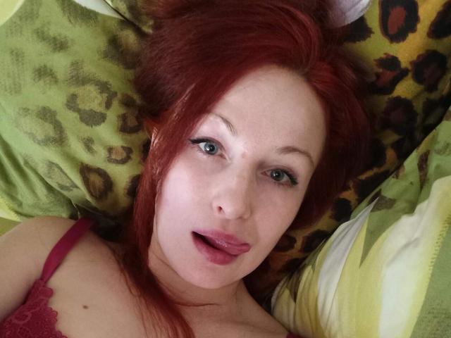 Marlene Davies, con i capelli rossi, gli occhi verdi e il rossetto rosa, la lingua leggermente fuori, sdraiata su un cuscino a stampa leopardo, indossa un reggiseno di pizzo rosso, mostrando il seno..