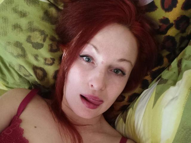 Marlene Davies, com cabelo ruivo, olhos verdes e lábios rosados, vestindo um sutiã de renda vermelho, deitada em um travesseiro com estampa de leopardo, com a língua ligeiramente para fora, olhando para a câmera..
