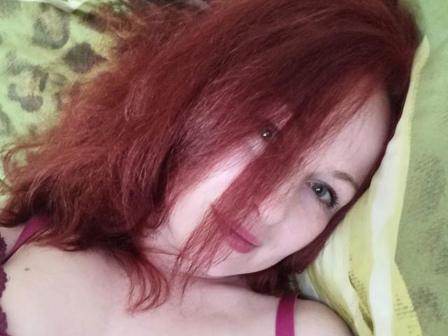 Marlene Davies, com cabelo ruivo, um pequeno piercing no nariz, olhos verdes e um sutiã roxo, deitada em um travesseiro amarelo, sorrindo levemente, mostrando seus seios..