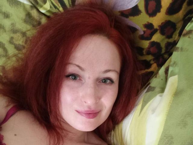MarleneDavies has red hair, green eyes, and a small smile. She's lying on a leopard-print pillow with green and white patterns, wearing a pink strap.