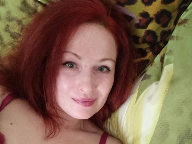 Marlene Davies, avec ses cheveux roux, ses yeux verts et son rouge à lèvres rose, sourit à l'appareil photo, allongée sur un oreiller vert et jaune. Ses petites seins sont visibles sous un soutien-gorge rouge..