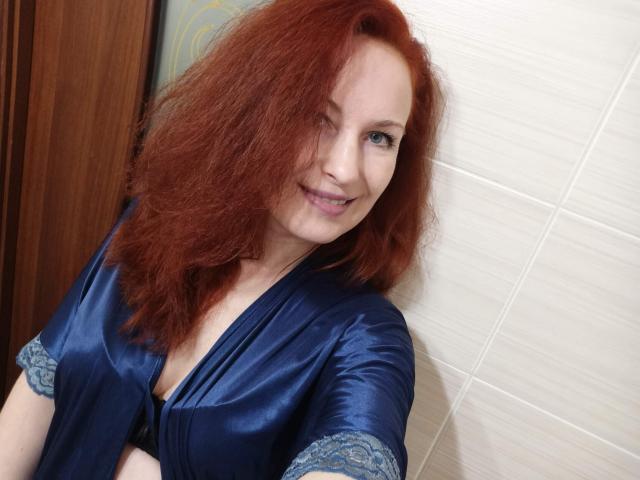 MarleneDavies with red curly hair, smiling, wearing a blue satin robe with lace cuffs, standing against a tiled wall, showing her small tits and a cute face.