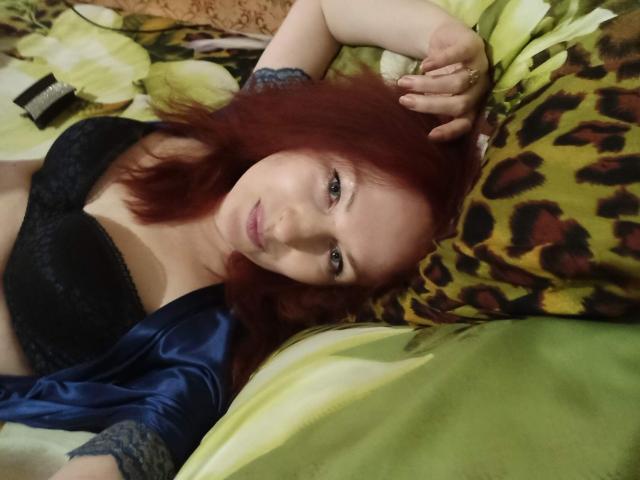 Marlene Davies, com cabelo ruivo, sutiã de renda preta e roupão de cetim azul, deitada em uma cama com estampas verdes e de leopardo, com a mão no cabelo, olhando para a câmera, mostrando seus seios..
