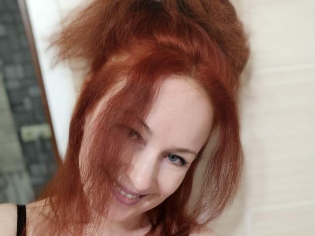 Marlene Davies, avec ses cheveux roux en bataille, souriante, aux yeux bleus, à la peau claire, portant un haut noir, et avec un regard mignon et légèrement excentrique, appuyée contre une porte blanche..