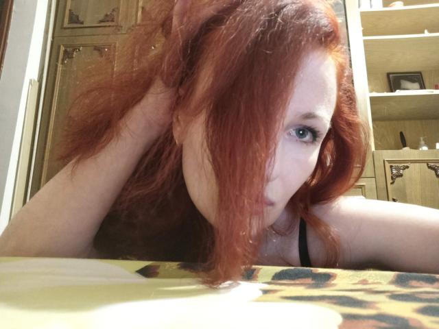 MarleneDavies with red hair, blue eyes, black tank top, leaning on table, hand in hair, looking at camera, pale skin, slightly messy hair, yellow patterned table in foreground.