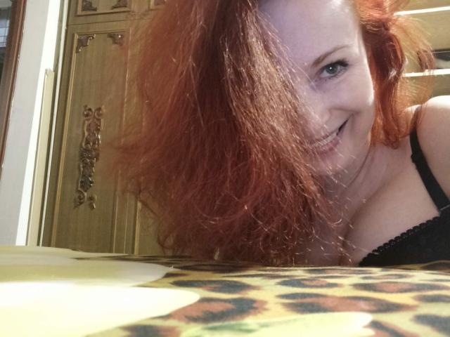 Marlene Davies, con cabello rojo, sonriendo a la cámara, vistiendo un sujetador de encaje negro, mostrando sus senos, inclinándose hacia adelante, sobre una manta de estampado de leopardo, con puertas de madera en el fondo..