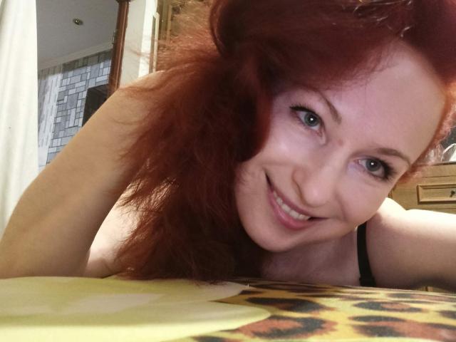 MarleneDavies, red-haired woman, smiling, lying on bed, black strap visible, looking at camera, brown eyes, fair skin, messy hair, yellow and leopard print blanket in foreground.