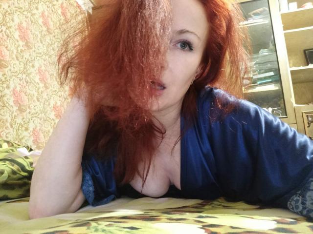 Marlene Davies, con cabello rojo, camisa azul y senos visibles, está acostada en una cama, mirando a la cámara con la boca ligeramente abierta. Su cabello cubre un ojo..
