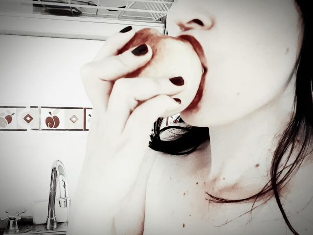 AliceRossi with black nail polish eating an apple, her mouth full, upper chest and neck visible, slight blush on pale skin, brown hair partially seen, in a kitchen.