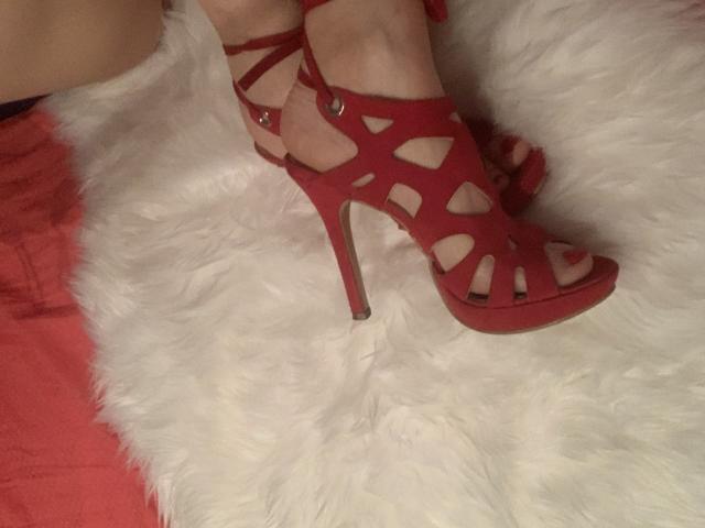 RedheadRita69's foot in a red, high-heel, cut-out sandal with ankle straps, resting on a white, fluffy, textured surface. Her toes are painted red.