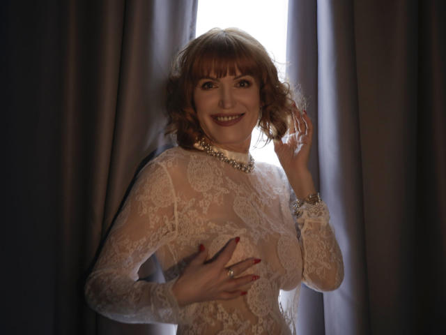 RedheadRita69 met krullend rood haar, draagt een doorschijnende kantjurk, glimlacht, met haar hand op haar borst, een ketting, een ring en rode nagels, en staat voor een helder raam..