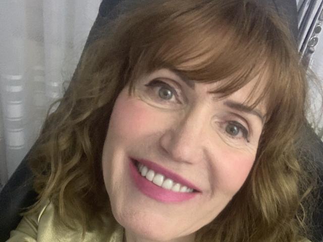 RedheadRita69 has wavy brown hair, green eyes, pink lipstick, and a smiling face with visible teeth. She's wearing a gold top, looking happy and cute.