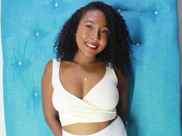 CaroldelaTorre, met krullend zwart haar, lachend, draagt een witte crop top die haar borsten laat zien, met op de achtergrond een blauwe, pluizige achtergrond. Ze heeft een gouden ketting om en rode lippenstift op..