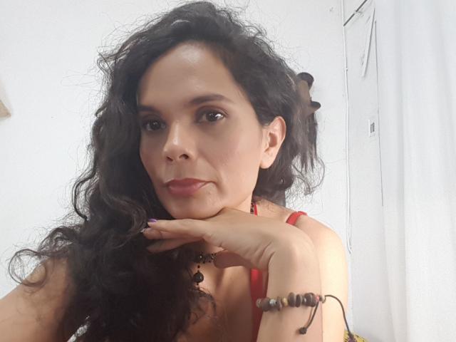 Natthally has curly black hair, medium tits, and a red tank top. She's resting her chin on her hand, with a serious expression and red lipstick.