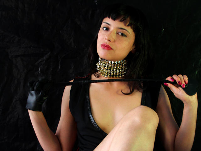 Marlaa with black bangs, wearing a spiked choker, black dress, and gloves, holding a whip, sitting with one knee up, showing her tits and pink nail polish.