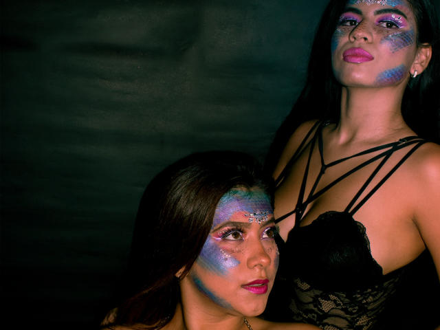 KorinYAmbar has blue glitter on her face, wearing black lace, with pink lipstick. Another KorinYAmbar, with similar face glitter, looks up at her with dark hair.