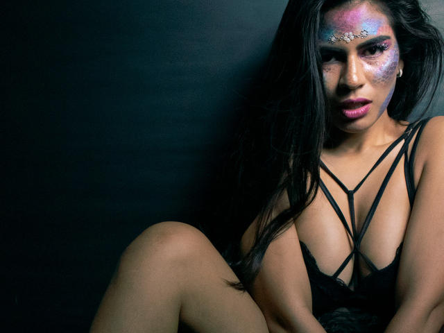 KorinYAmbar tiene pintura facial de colores, lencería negra con tirantes finos y cabello negro largo, está sentada con las piernas cruzadas, mostrando sus pechos y con una mirada seductora, sobre un fondo oscuro..