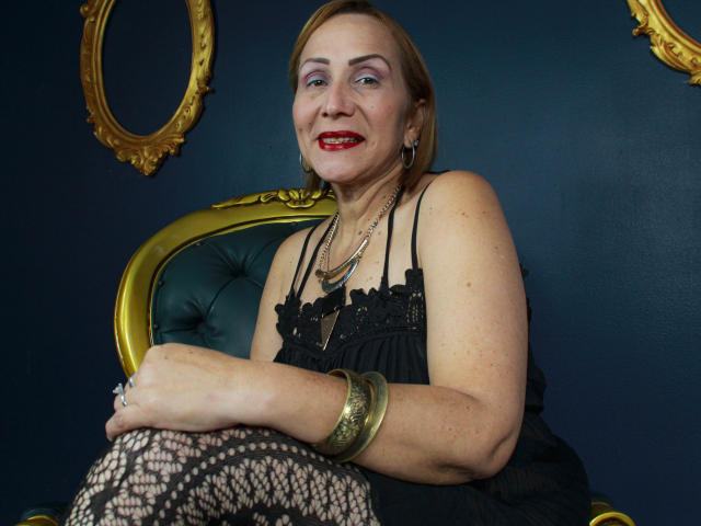 DianeOasis con lápiz labial rojo, vestido de encaje negro, pulsera dorada y medias de rejilla, sentada en una silla verde y dorada, sonriendo. Tiene el cabello rubio corto y sombra de ojos morada..