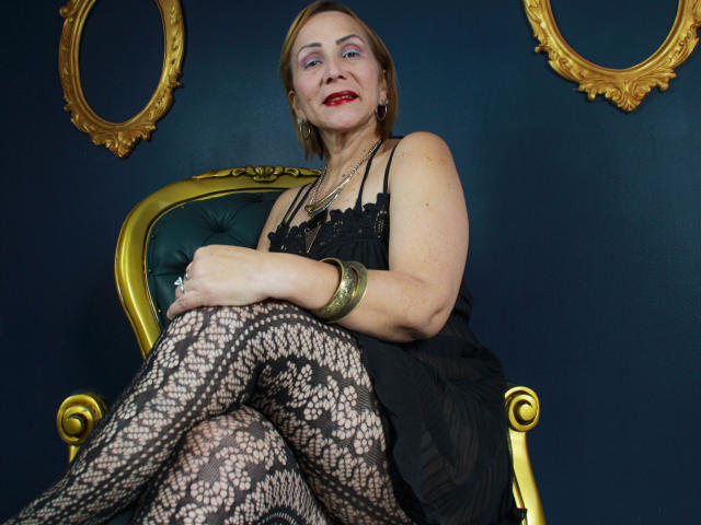 DianeOasis, una donna di mezza età con rossetto rosso, un abito di pizzo nero e collant a rete, seduta su una sedia dorata, che indossa un braccialetto verde, sorridente, con le gambe incrociate..