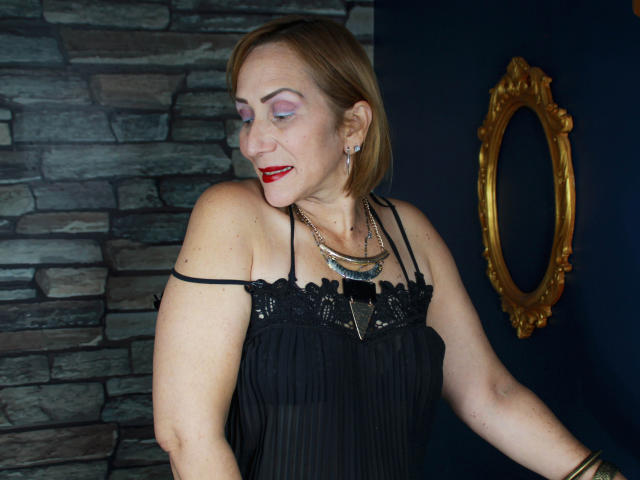 DianeOasis avec du rouge à lèvres rouge, des cheveux blonds courts, une robe en dentelle noire, un collier en argent, du fard à paupières violet et du rouge à lèvres rouge, montrant légèrement ses seins, debout devant un mur sombre..