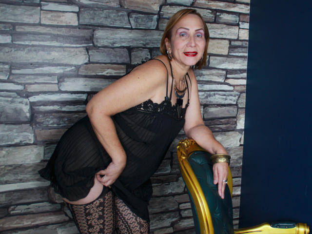 DianeOasis, middle-aged woman with red lipstick, black lace lingerie, and patterned stockings, bending over a golden chair, showing her black lace-covered ass and lace-patterned thighs.