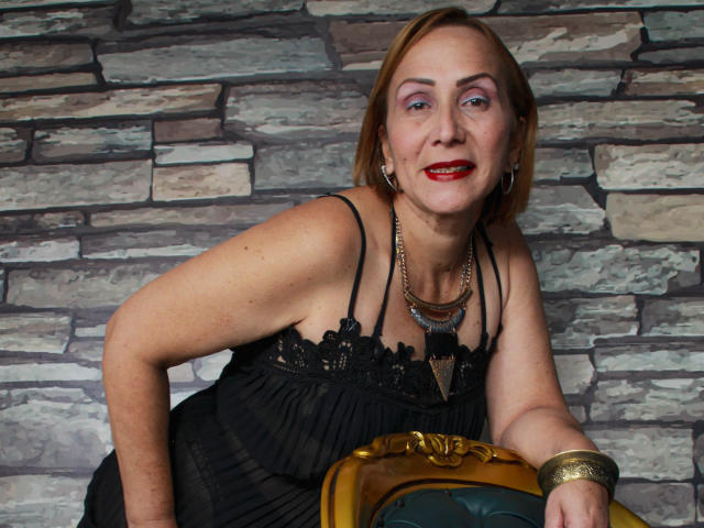 DianeOasis has red lipstick, purple eye makeup, black lace dress, gold bracelet, necklace, leaning on a yellow chair with her arm raised, smiling, short blonde hair.