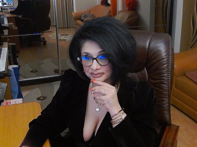 ClassybutNaughty, met blauwe ogen, een zwarte bril en kort zwart haar, gekleed in een zwarte outfit, heeft haar vinger in de buurt van haar mond, toont haar borsten, en zit in een leren stoel..