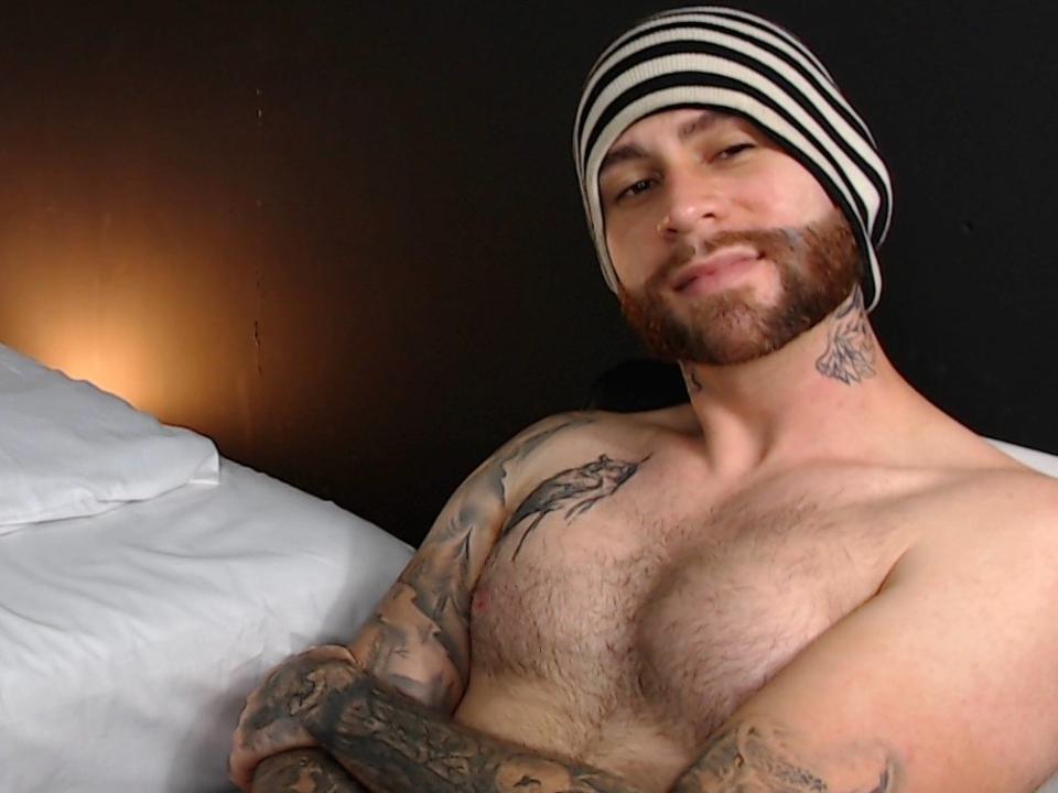 ZeusBrioly, sem camisa, com tatuagens, barbudo, usando um gorro listrado preto e branco, sorrindo, com os braços cruzados, peito peludo, cama com travesseiros brancos ao fundo..