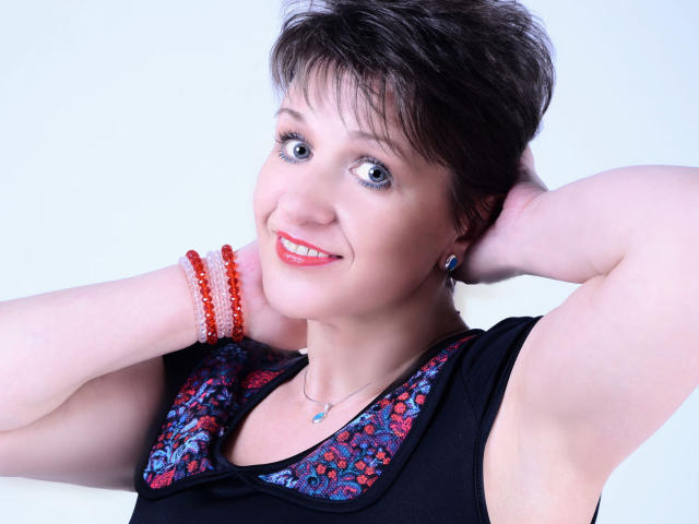 DoraBlueEyess with short brown hair, blue eyes, and red lipstick, smiling, arms raised, wearing a black top with red and blue floral patterns, and red beaded bracelets.