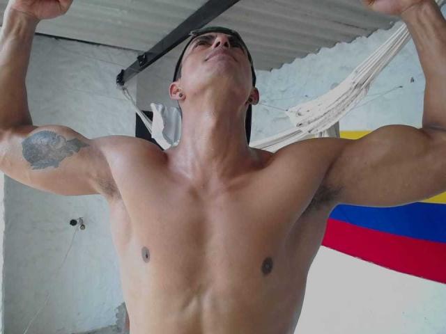 VictorHotX, sem camisa, musculoso, braços levantados, tatuagem no bíceps direito, olhando para cima, sob uma rede branca, bandeira colombiana visível, cabelo preto curto, tom de pele médio, abdômen definido..