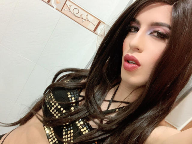 TiffanyTaylorHot has long brown hair, pink eyeshadow, red lipstick, and is wearing a black, gold-studded bra, showing her tits and a sexy, open-mouthed, confident expression.