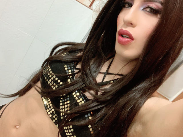 Tiffany Taylor, atractiva, con cabello largo y castaño, vistiendo un sujetador negro con tachuelas doradas, mostrando sus senos, con lápiz labial rosa y maquillaje de ojos morado, tomándose un selfie..