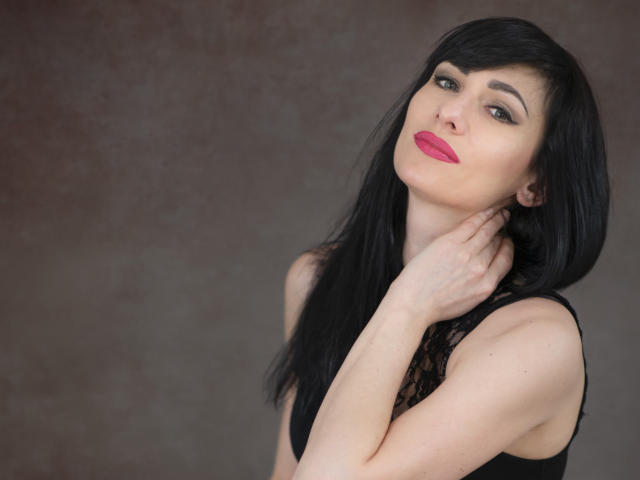 HypnoticIsabel with black hair, red lipstick, and black lace top, holding her neck, showing her ass, and looking at the camera with a slight smile.