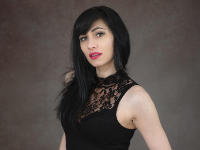 HypnoticIsabel with black lace dress, red lipstick, and straight black hair, showing off her tits and confident gaze, against a muted background.