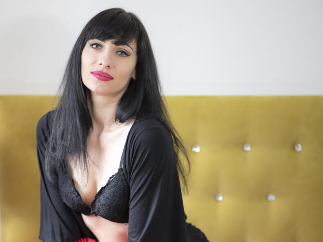 HypnoticIsabel with black lace bra, showing her boobs, has long black hair, bangs, pink lipstick, and black robe, sitting on yellow tufted bed.