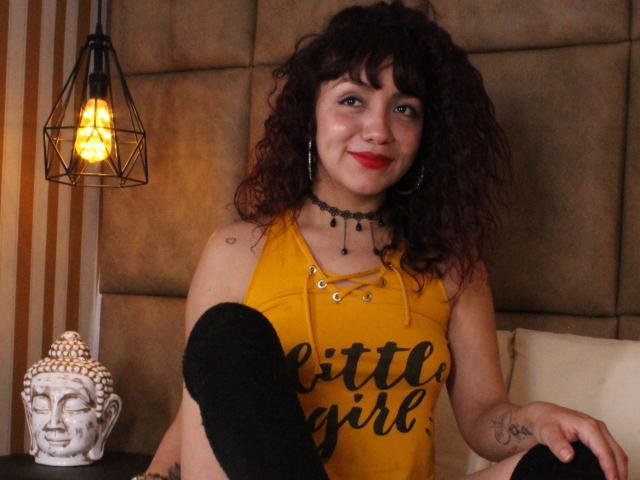 HottyKendall with curly brown hair, red lipstick, yellow sleeveless top, black choker, black socks, and tattoos, smiling at the camera. Her tits are visible. Lamp and Buddha statue in background.