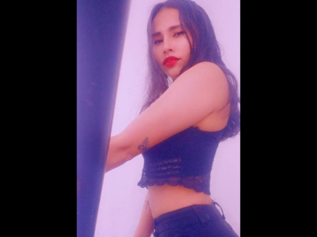 KarolineLovely, com cabelos longos e escuros, batom vermelho, cropped top de renda preta e jeans pretos, exibindo uma tatuagem no braço direito, está em pé ao lado de uma cortina escura..