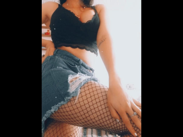 KarolineLovely mit gebräunter Haut, schwarzem Spitzenoberteil, Jeansshorts, zerrissen, Fischernetzstrümpfen, die ihre Brüste und ihren Hintern zeigen, Tattoo am linken Arm, Hand auf dem Oberschenkel, heller Hintergrund..