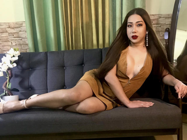 Eine verführerische Puppe mit langen, braunen Haaren, die ein braunes Kleid mit tiefem Ausschnitt trägt, liegt auf einem grauen Sofa und zeigt ihre Brüste, Beine und roten Lippen, und trägt weiße High Heels..
