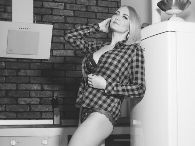 Black-and-white photo of EliseONE with blonde hair, wearing an unbuttoned plaid shirt, exposing her large tits and lace panties, leaning against a fridge in a kitchen.
