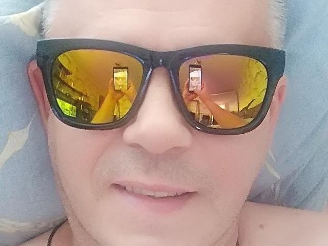 ArthurXSky met gele reflecterende zonnebril, die een telefoon vasthoudt, zichtbaar in de reflectie, liggend, bleke huid, lichte baardgroei, lichte glimlach, blauw kussen, kort wit haar, zwarte montuur bril..