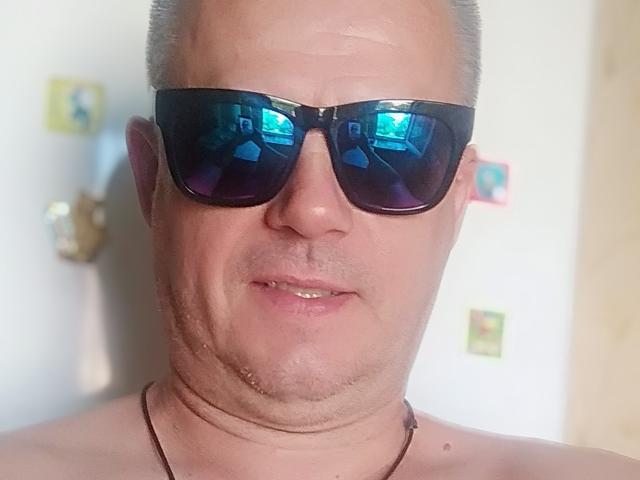 ArthurXSky, met een kaal hoofd, draagt zwarte zonnebrillen die blauw licht reflecteren, heeft lichte stoppels, is naakt boven en draagt een zwarte ketting, met een neutrale gezichtsuitdrukking..