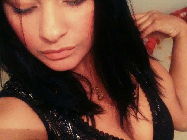 KarlysHoney trägt einen schwarzen Spitzen-BH, auf dem ihre Brüste zu sehen sind. Sie hat dunkles Haar und einen leichten Schmollmund. Sie trägt eine Halskette und ist auf einem leicht unscharfen Selfie zu sehen..