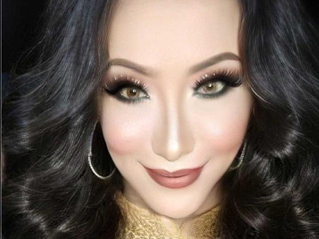 WOWPinayWilFrid hat dunkles, welliges Haar, grüne Augen mit starkem schwarzen Eyeliner und ein leichtes Lächeln mit nuder Farblippenstift. Sie trägt große silberne Ohrringe im Stecker-Stil und ein Oberteil mit goldenem Muster..
