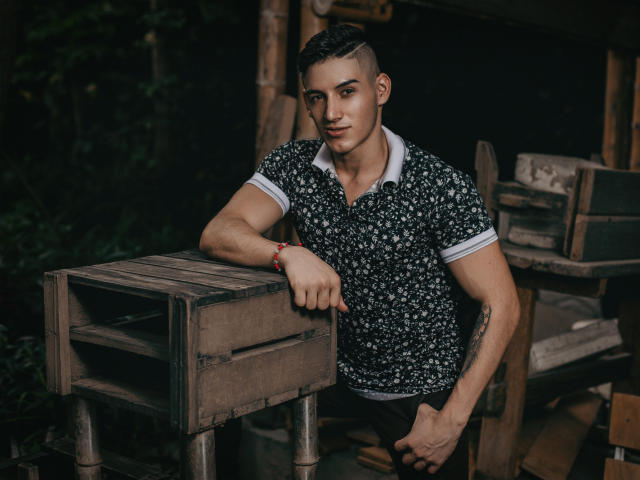 JonathanJeremiah with a muscular build, short dark hair, and a floral shirt, leaning on a wooden crate, wearing a red bracelet, looking at the camera with a slight smile.