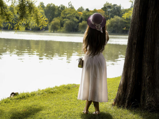 DashingFoxyX stands by a tree, wearing a white dress and purple hat, holding a small purse, with long brown hair, facing a calm lake. Ass is not visible.