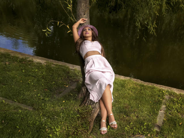 DashingFoxyX is leaning against a tree, wearing a purple hat, pink crop top, and pink skirt, with white sandals, her arms above her head, and her legs crossed.