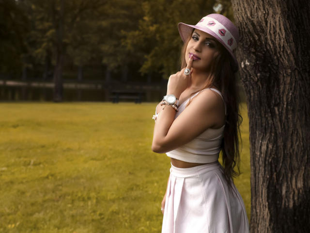 DashingFoxyX, con cabello castaño largo, un sombrero rosa, una camiseta blanca y una falda rosa, está de pie junto a un árbol, con un dedo en los labios, luciendo sexy con su trasero ligeramente visible..